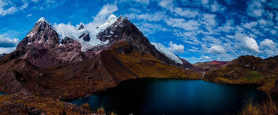 Panoramic view of snow-capped mountains with a lake at their base under a blue sky.