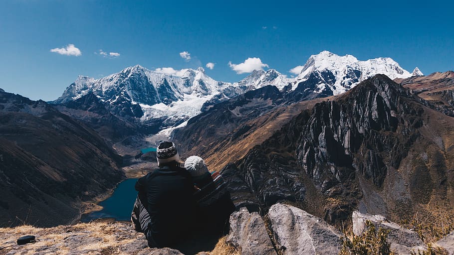 Person sitting on a mountain overlooking a valley with snow-capped mountains.