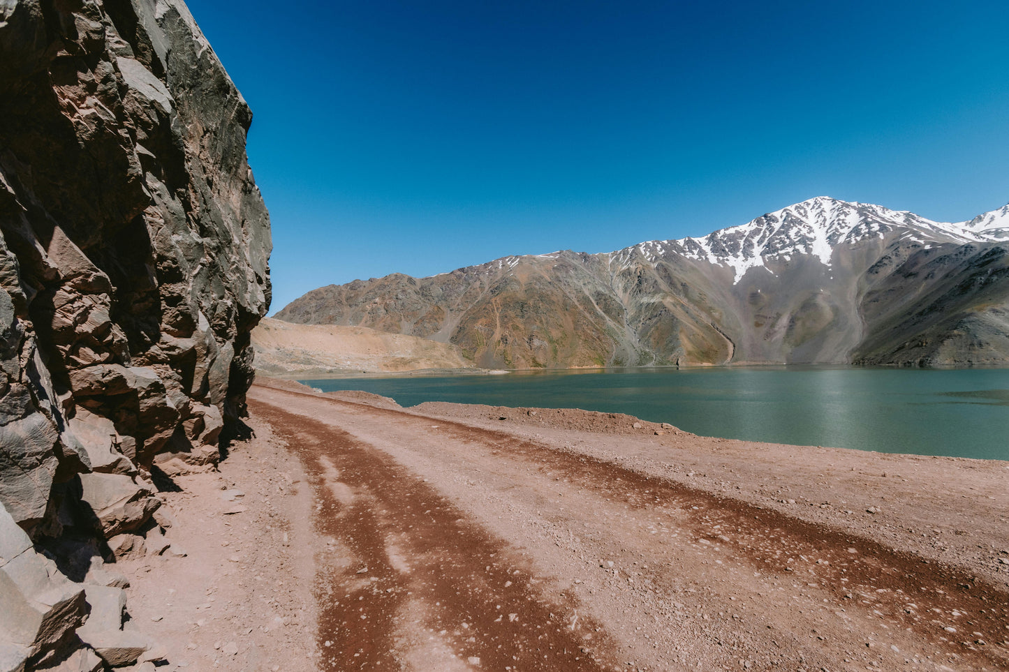 Mountain lake with a dirt path leading to it, surrounded by rocky terrain and snow-capped mountains.