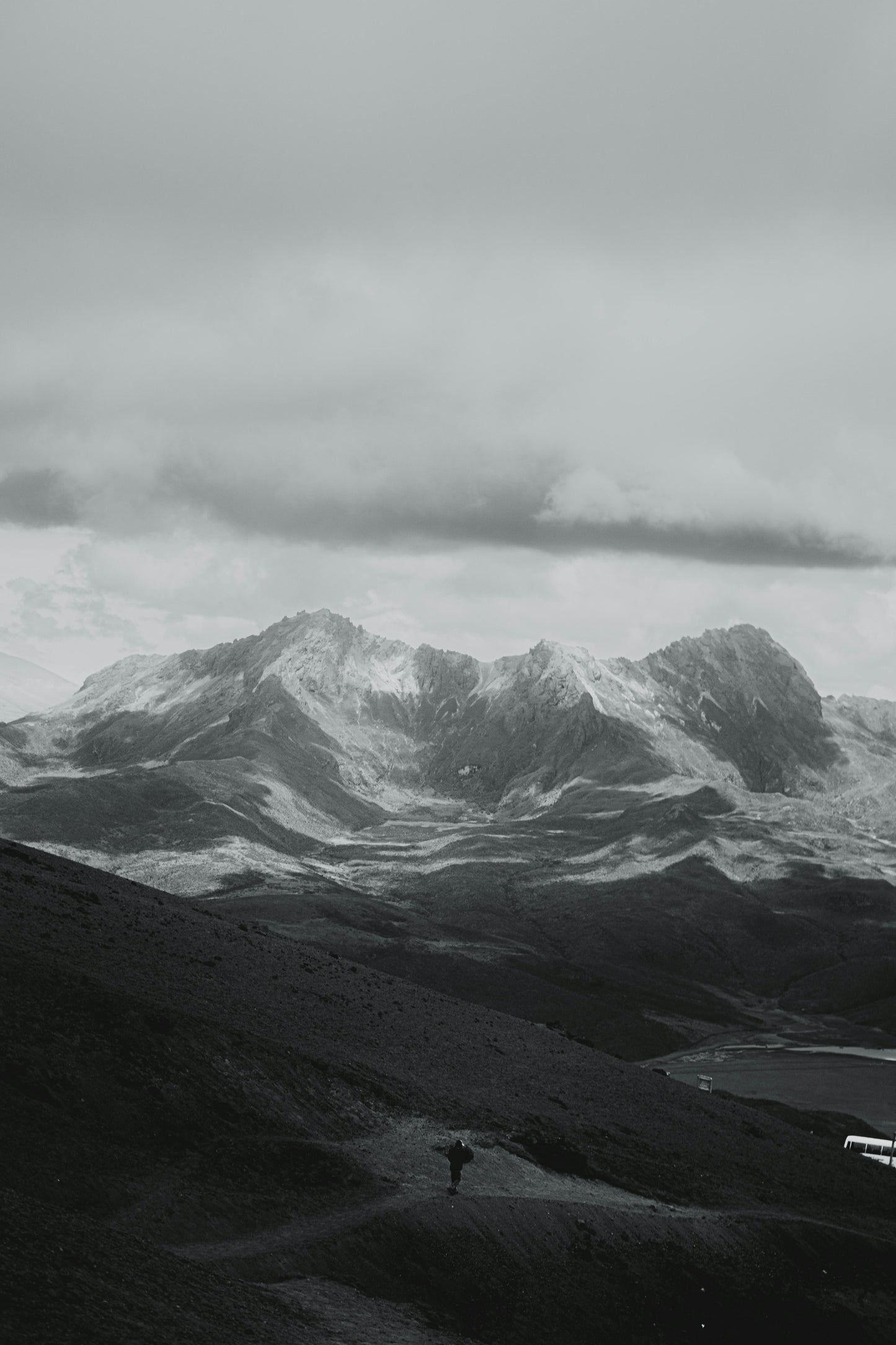 Person standing on a mountain path with snow-capped peaks and clouds in the background