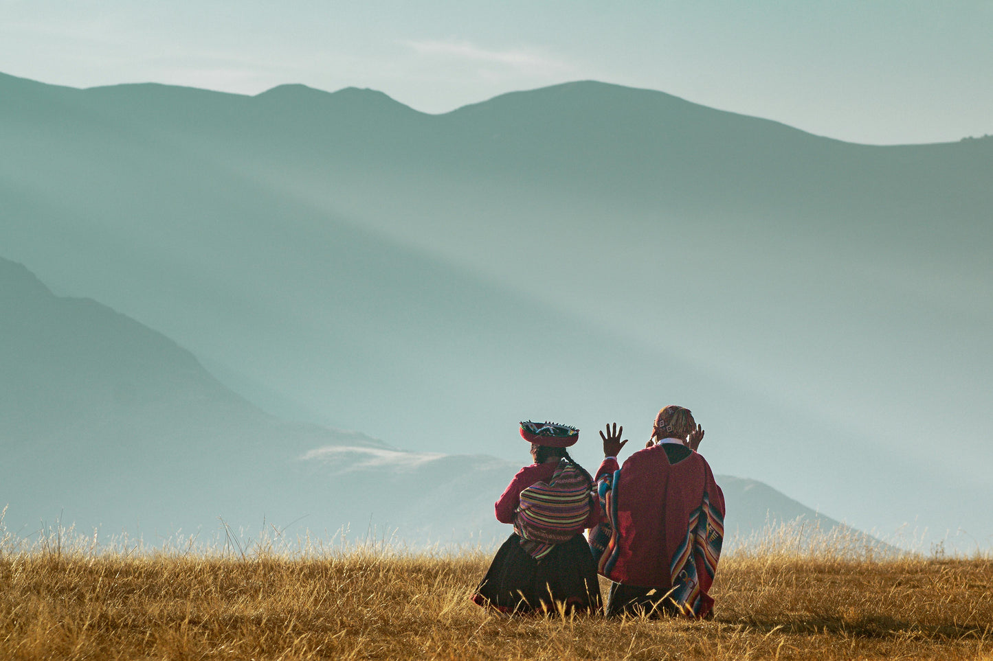 Two people sitting on a grassy hill with mountains in the background