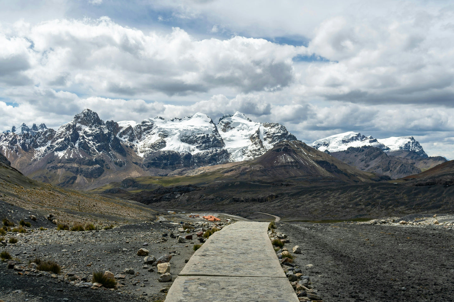Scenic view of a mountain range with a wooden path leading towards snow-capped peaks under a cloudy sky.