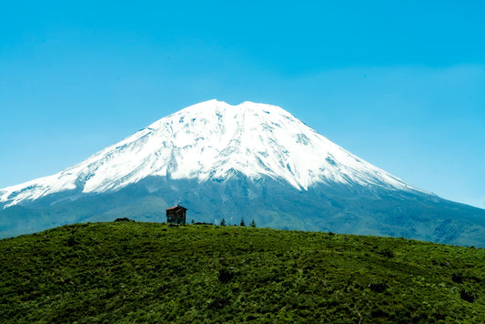 Snow-capped mountain with a small building on a grassy hill under a clear blue sky.