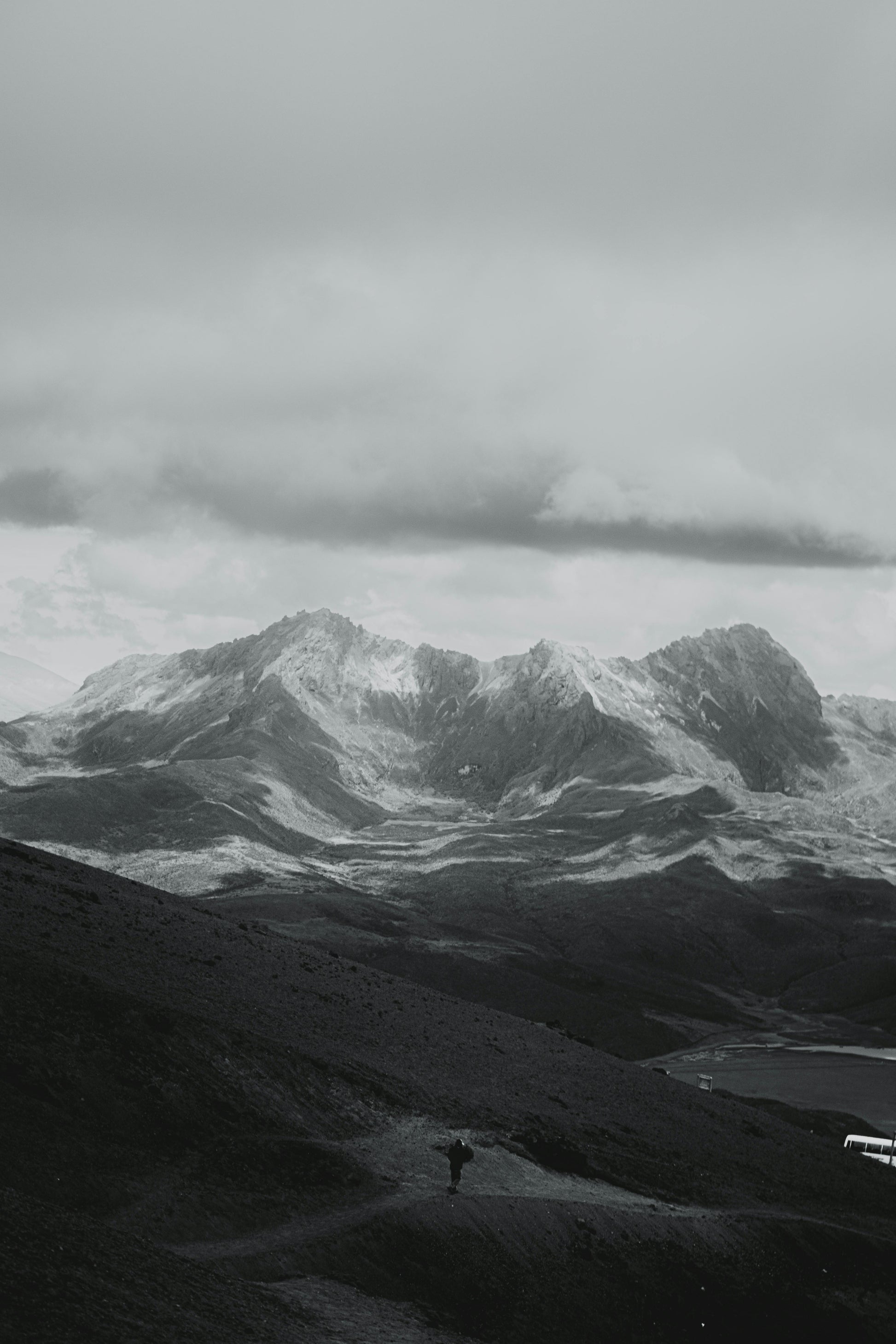 Person standing on a mountain path with snow-capped peaks and clouds in the background