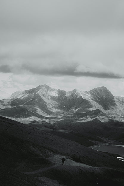 Person standing on a mountain path with snow-capped peaks and clouds in the background