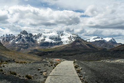 Scenic view of a mountain range with a wooden path leading towards snow-capped peaks under a cloudy sky.