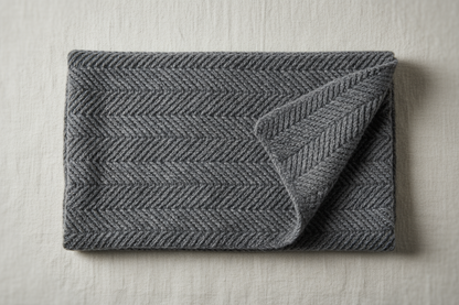 Flat lay image of a folded Herringbone Relief Knit Scarf in charcoal gray, placed neatly on a neutral off-white linen surface. Include a soft side light from the top-left corner to highlight the herringbone diagonal texture and subtle matte halo typical of baby alpaca fibers. Composition: one scarf folded in thirds, one corner turned slightly to reveal reverse side. Camera: top-down 90° angle, 50mm equivalent, soft focus edges. Texture should show tight uniform valleys and raised diagonal lines.